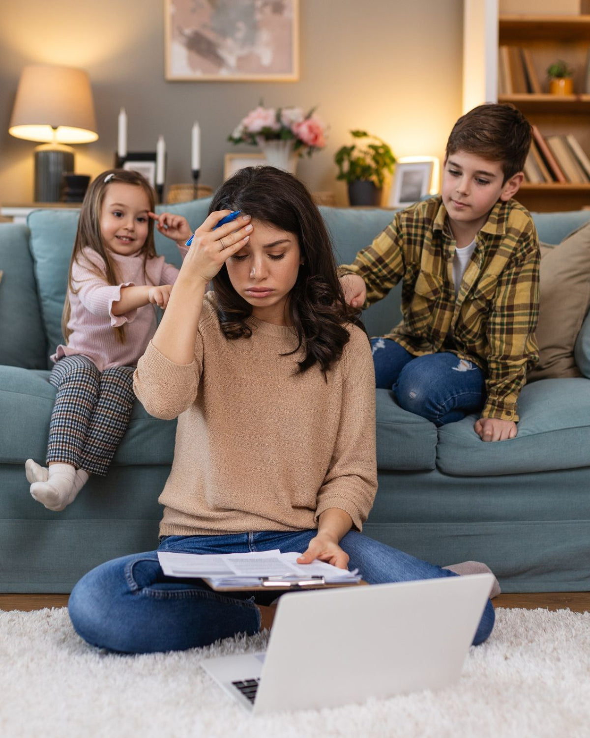 Woman sitting on the floor with a laptop, surrounded by two children on a couch.
